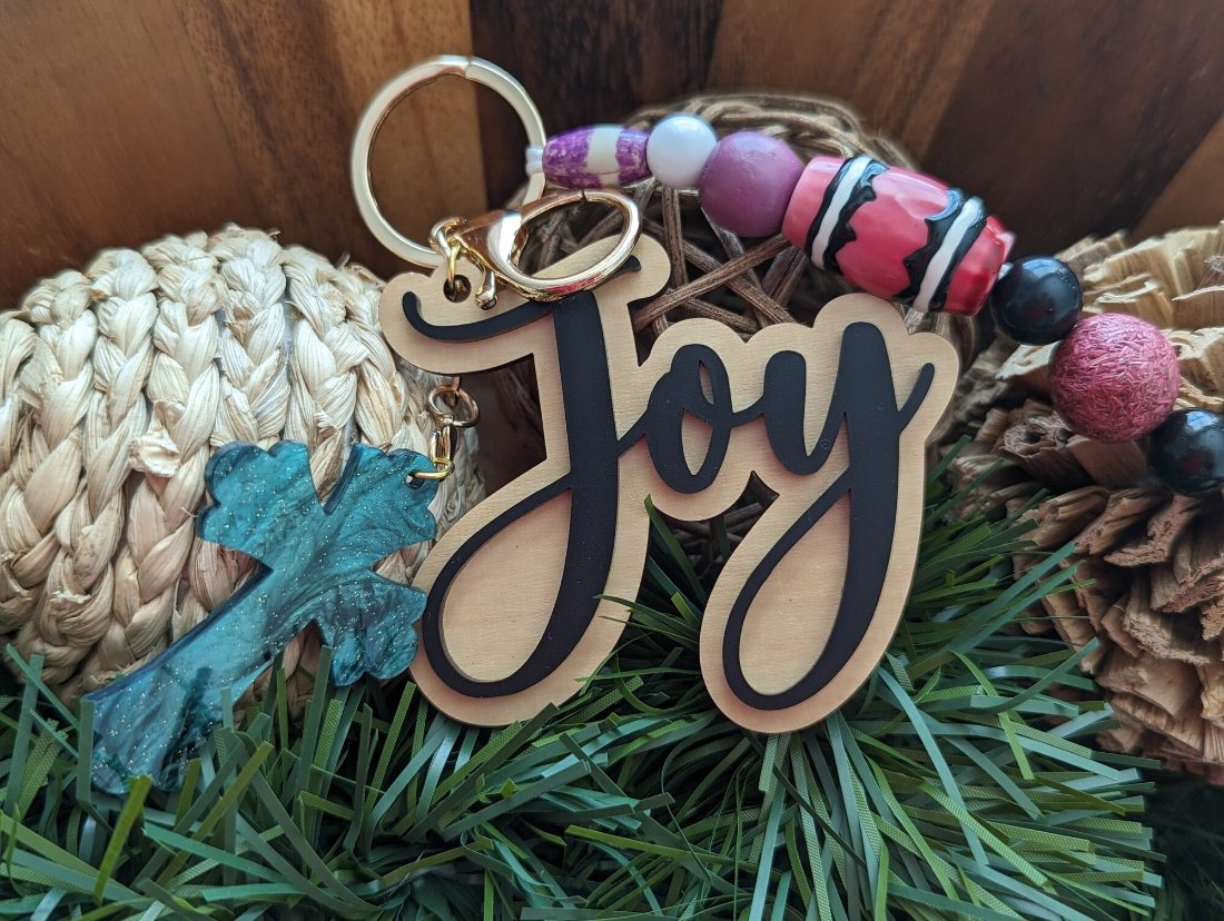 A wood and black acrylic ‘Joy’ charm with pink beads and a teal resin cross nestled in greenery
